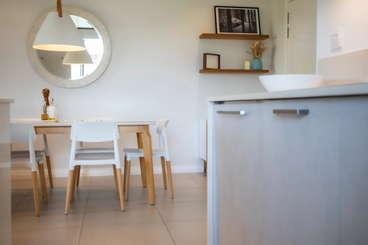 Espace salle à manger lumineux avec table en bois et chaises blanches. Miroir rond et étagères décoratives ajoutent une touche moderne.