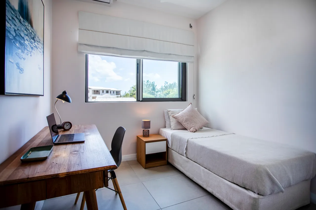 Sol en carrelage clair et mobilier en bois pour une ambiance simple. Bureau en bois avec lampe et chaise noire, offrant un espace de travail. Fenêtre laissant entrer la lumière naturelle dans la pièce.