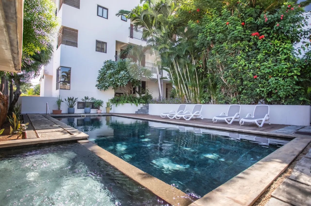 Teintes vertes dominent l'extérieur, créant une ambiance naturelle. La piscine est encadrée par des plantes tropicales. Chaises longues blanches disposent d'un espace ombragé. Mur en pierre ajoute un contraste aux espaces extérieurs. Accès direct depuis la galerie pour profiter de l'extérieur.