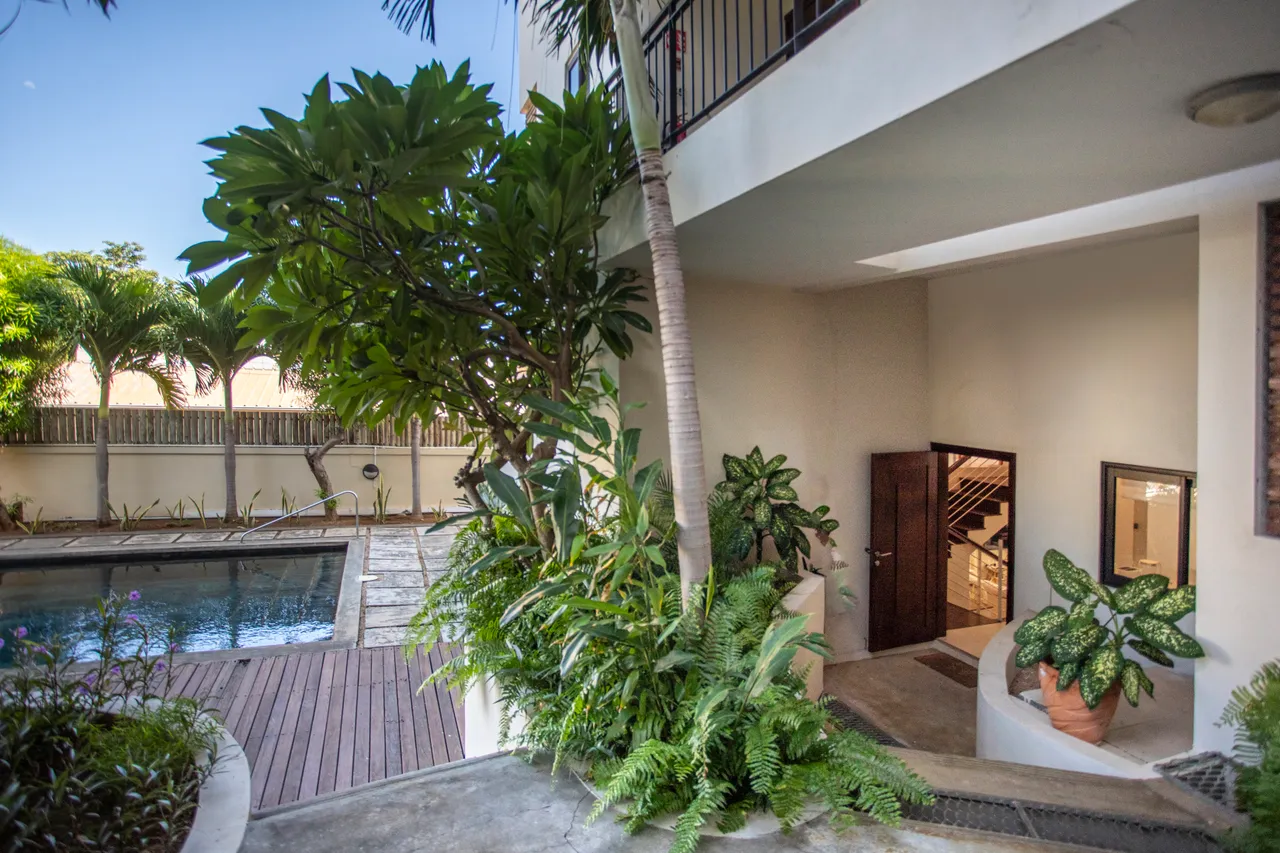 Galerie. Accès direct à la piscine depuis l'intérieur avec verdure environnante.  

Escaliers menant à l'étage, éclairés par la lumière naturelle.  

Plantes tropicales ajoutent une touche de verdure à l'entrée.  

Mur en pierre délimite l'espace, offrant un contraste avec la piscine.  

La zone autour de la piscine est accessible et soigneusement