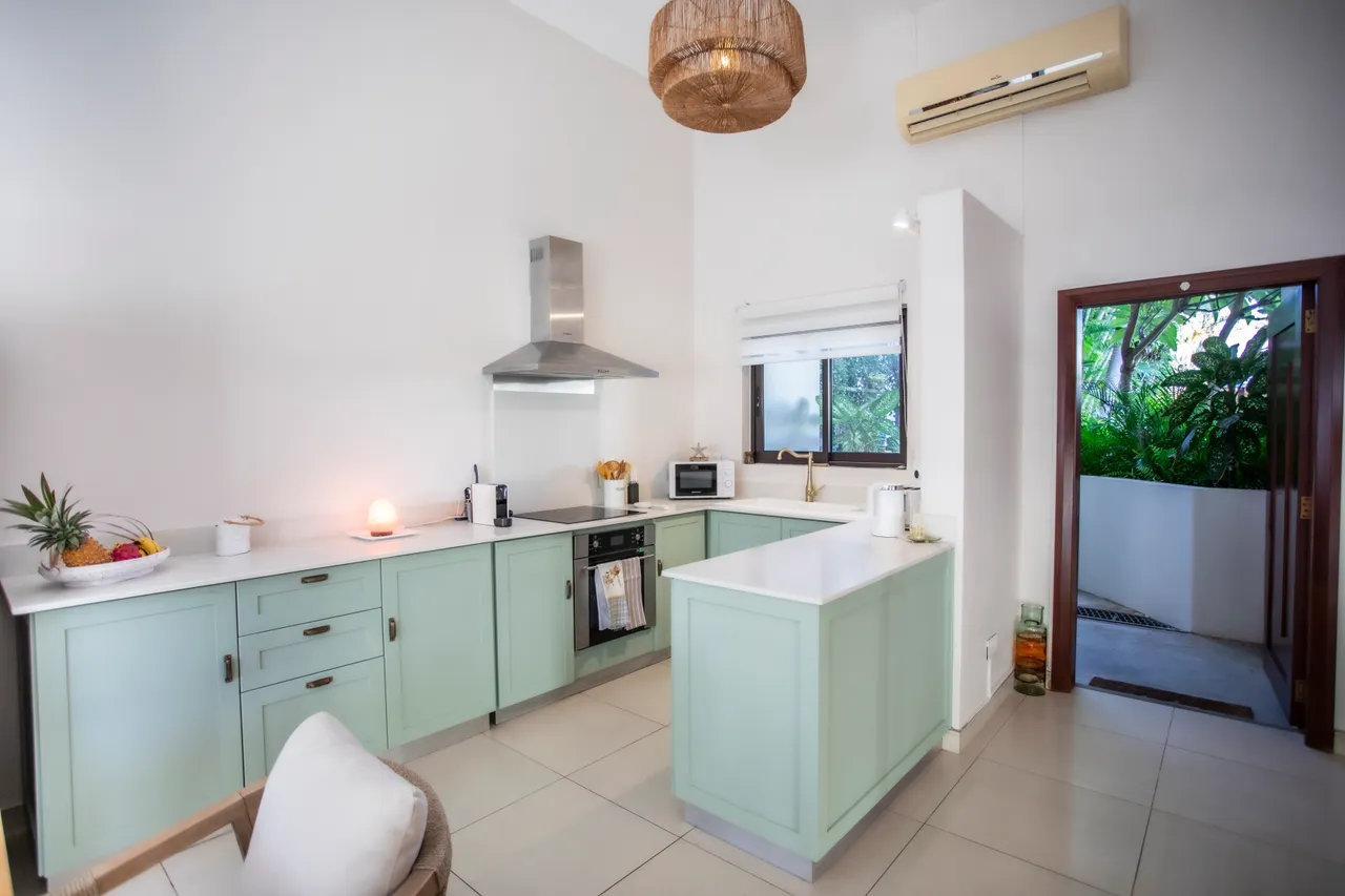 Cuisine moderne avec des meubles en bois clair et un plan de travail spacieux.  

Équipements récents incluent un four et un réfrigérateur encastré.  

Lumière naturelle entre par la fenêtre donnant sur la verdure environnante.  

L'îlot central sert de surface de préparation et de coin repas.  

Détails en pastel apportent une note de fraîcheur à