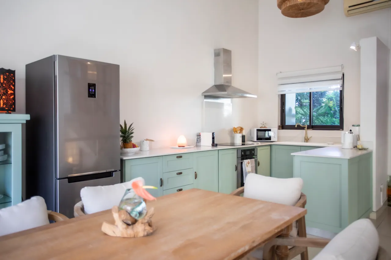 Cuisine avec meubles en bois clair et équipements modernes.  

Table en bois entourée de chaises confortables pour les repas.  

Fenêtre laisse entrer la lumière, offrant une vue sur le jardin.  

Électroménagers récents, dont un réfrigérateur et un four encastré.  

L'îlot central sert à la préparation et à la convivialité.
