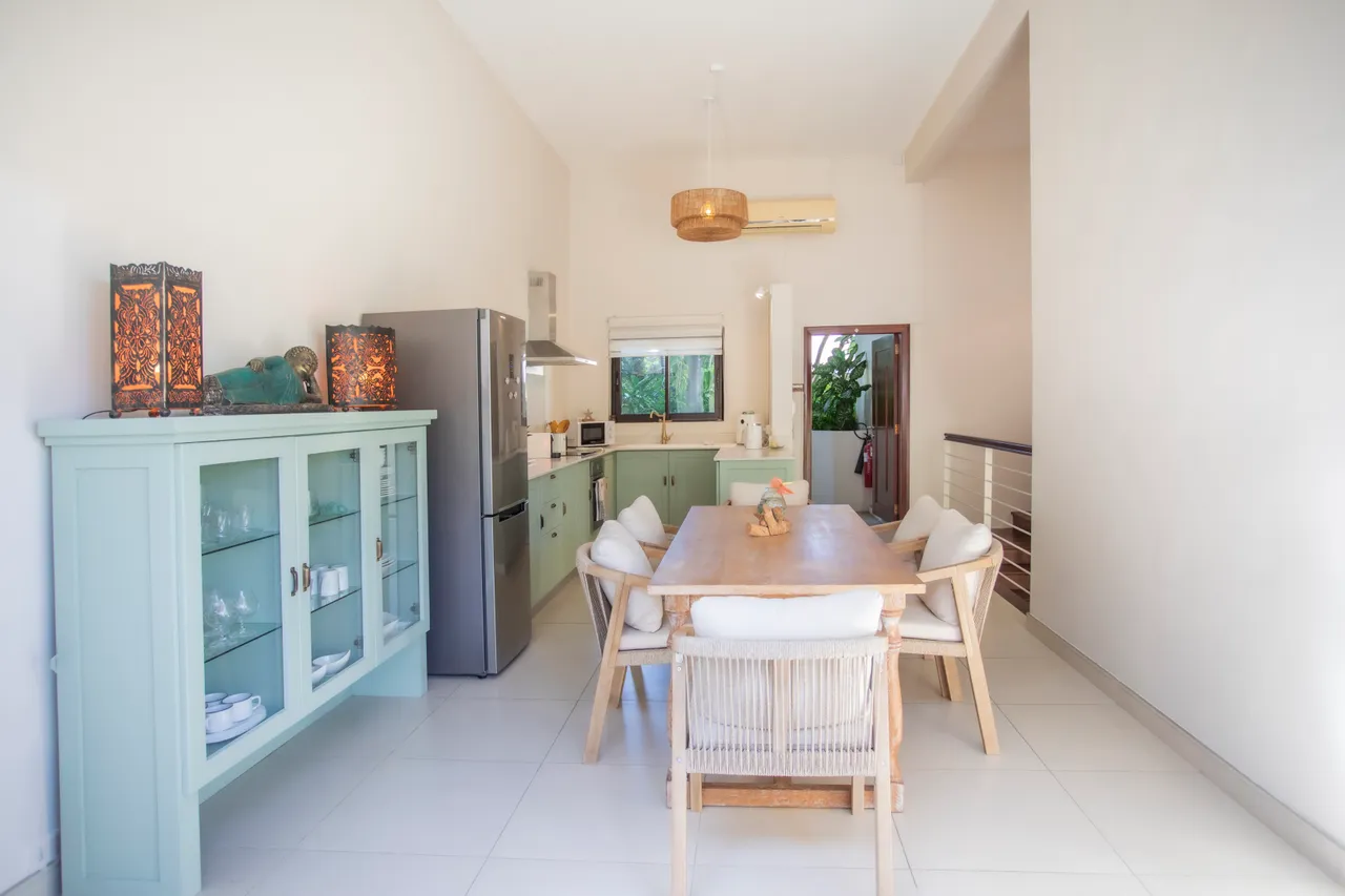 Espace salle à manger lumineux avec une grande table en bois et chaises confortables.  

Mobilier de rangement en bois présente des éléments décoratifs intégrés.  

Fenêtres offrent une vue sur la verdure, apportant une ambiance sereine.  

Électroménagers modernes, y compris un réfrigérateur en acier inoxydable.  

L'éclairage suspendu crée une atmos