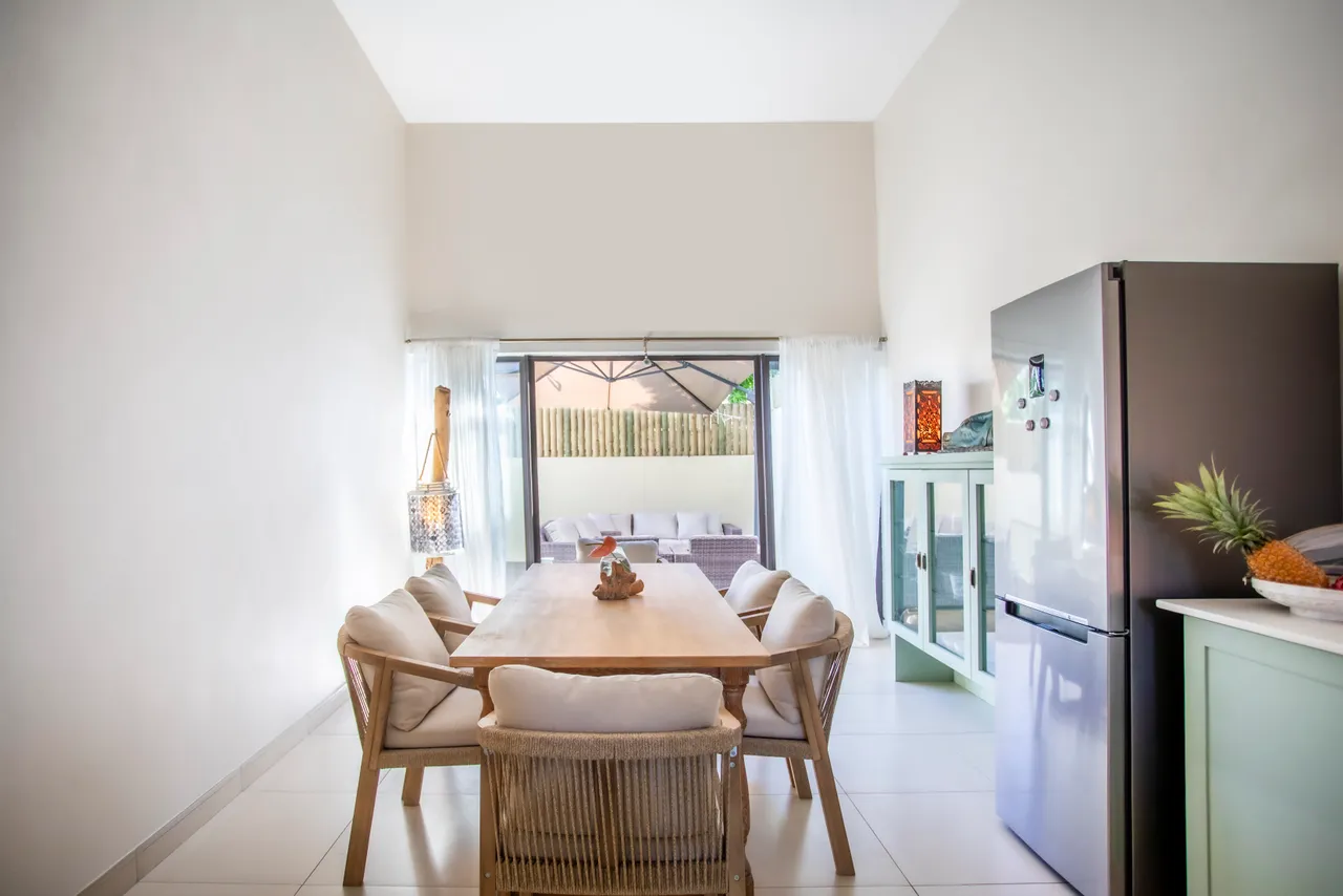 Espace salle à manger avec une grande table en bois et chaises confortables.  

Fenêtres ouvertes sur un patio, apportant lumière naturelle et ventilation.  

Électroménagers modernes, dont un réfrigérateur, intégrés au design.  

Détail en rotin sur les chaises ajoute une touche de texture.  

Ambiance calme grâce aux teintes claires des murs