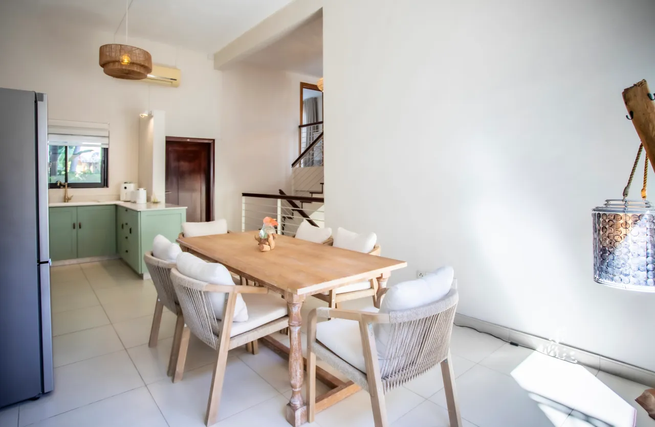 Table en bois avec chaises en rotin, créant un espace convivial.  

Fenêtre laisse entrer la lumière et offre une vue sur l'extérieur.  

Électroménagers intégrés, dont un réfrigérateur, pour une cuisine fonctionnelle.  

L'éclairage naturel met en valeur les teintes claires des murs.  

Détails en rotin sur les chaises ajoutent