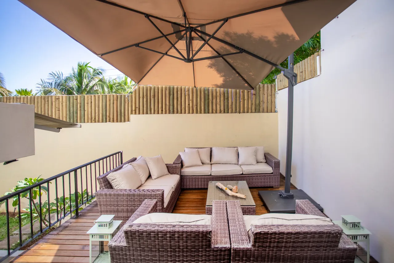Espace extérieur avec canapé en rotin et table basse en bois.  

Parasol offre de l'ombre pour des moments de détente.  

Plantes vertes en arrière-plan ajoutent une touche de nature.  

Sol en bois favorise une ambiance chaleureuse et accueillante.  

Coin salon confortable, idéal pour se relaxer en plein air.