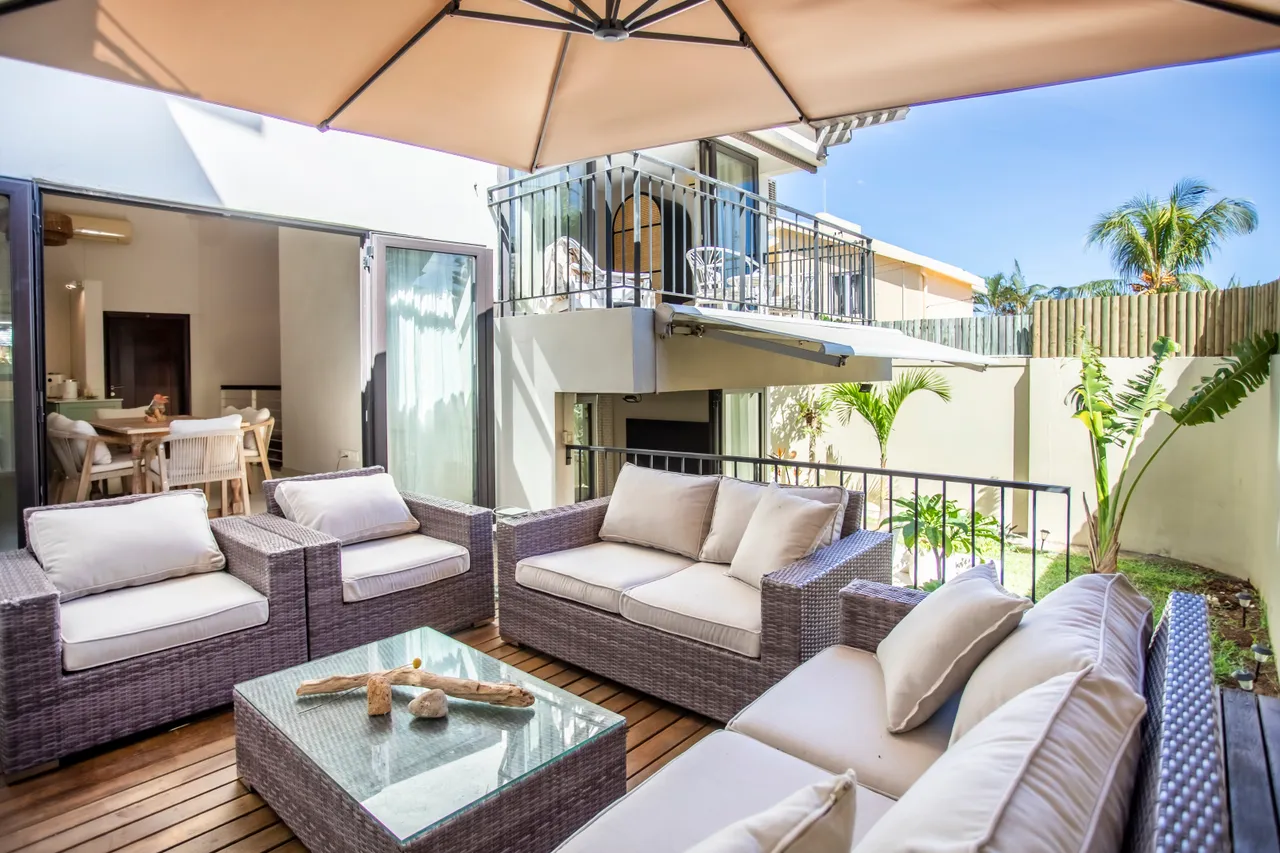 Espace extérieur avec canapé en rotin et table basse en verre.  

Parasol procure de l'ombre pour le confort des occupants.  

Plantes vertes en arrière-plan enrichissent l'environnement naturel.  

Dalle en bois offrant une atmosphère chaleureuse et accueillante.  

Arrangement soigné favorisant des moments de détente à l'extérieur.