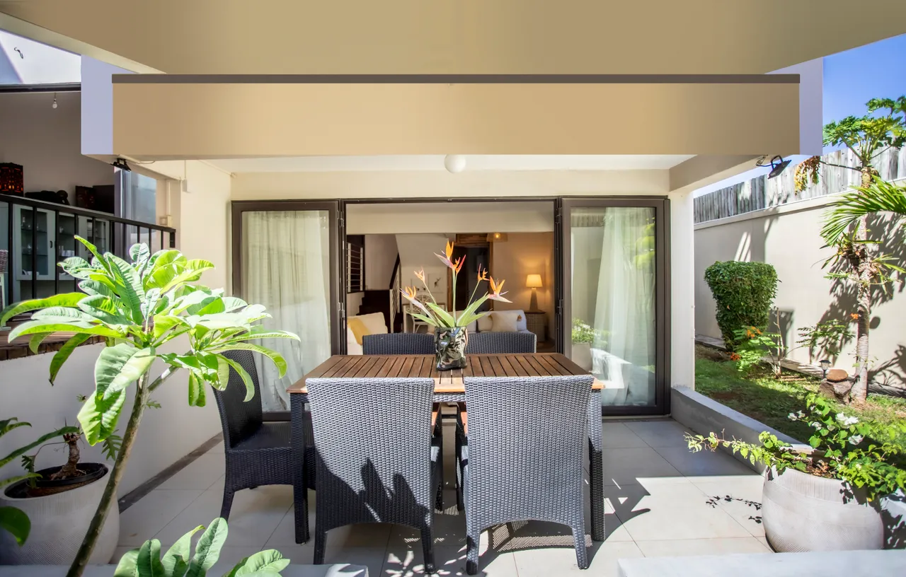 Espace extérieur avec table à manger en bois et chaises en rotin.  

Plantes décoratives ajoutent une touche naturelle à l'environnement.  

Accès à un coin salon confortable à proximité pour se détendre.  

Terrasse carrelée offrant une surface agréable pour les repas.  

Lumière naturelle éclaire l'espace tout au long de la journée.