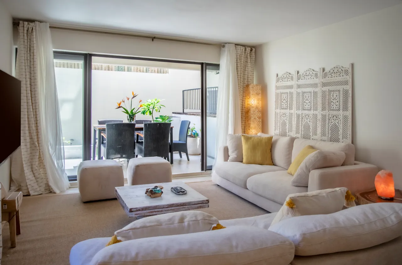 Espace de vie avec canapé clair et table basse en bois.  

Fenêtres à panneaux coulissants ouvrent sur la terrasse extérieure.  

Plantes en pot ajoutent une touche de verdure à l'intérieur.  

Éclairage indirect crée une ambiance douce et apaisante.  

Sol en jute offre une texture naturelle et chaleureuse.
