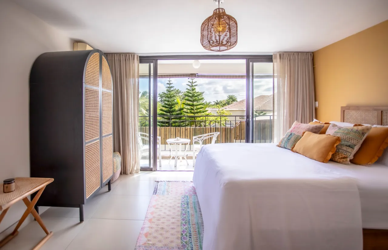 Tons clairs dominent la chambre, créant une atmosphère apaisante.  

Balcon privé accessible, offrant un espace extérieur supplémentaire.  

Linge de lit orné de coussins colorés apportant une touche de confort.  

Armoires en bois ajoutent du rangement tout en étant esthétiques.  

Luminaires suspendus diffusent une lumière douce dans l'espace.