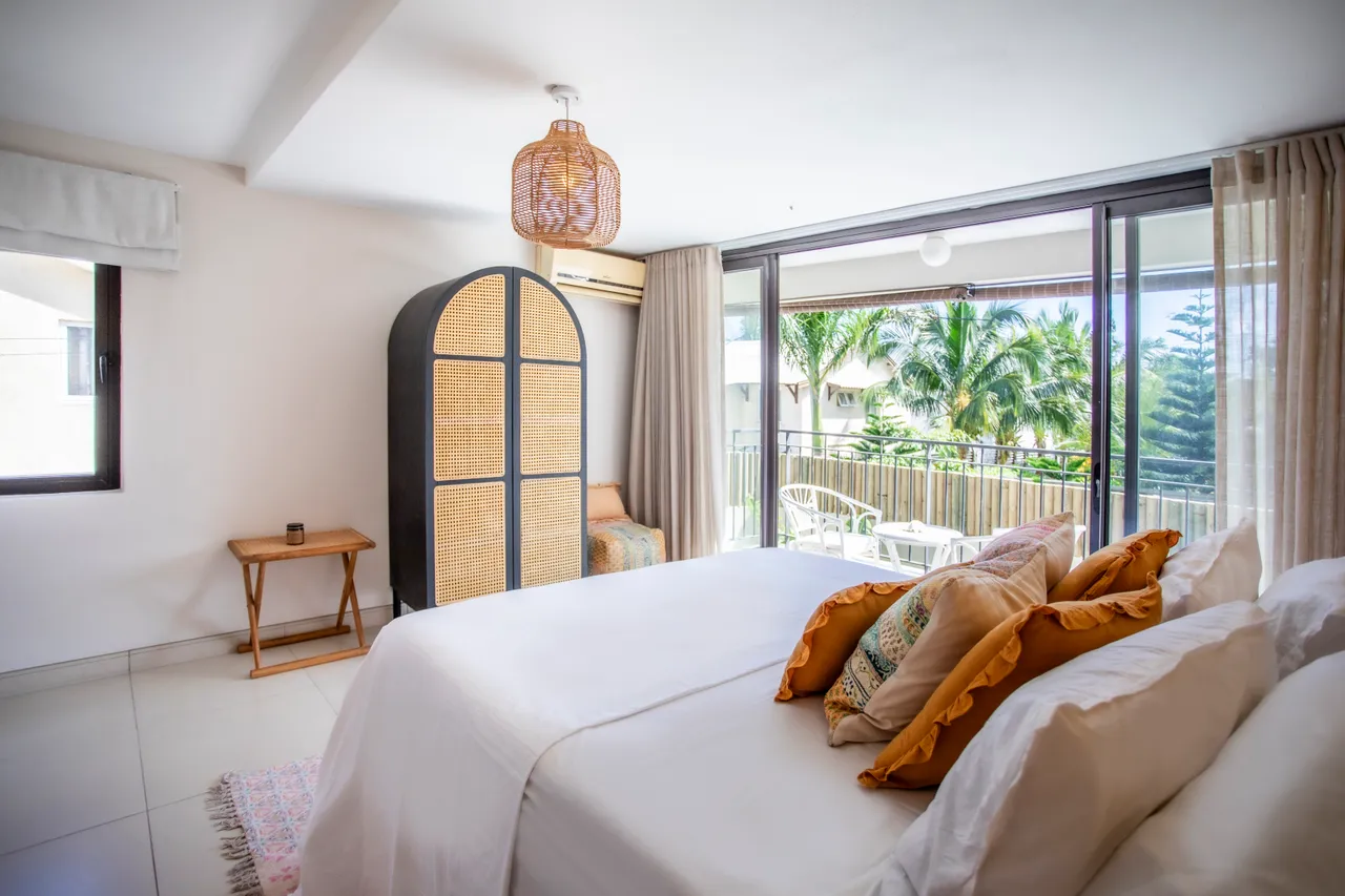 Chambre avec grandes fenêtres donnant sur un balcon privé.  

Linge de lit clair et coussins colorés ajoutent du confort.  

Mobilier en bois et éléments en osier créent une ambiance naturelle.  

Luminaire en rotin diffuse une lumière douce et chaleureuse.  

Espace de rangement intégré offrant fonctionnalité et esthétique.