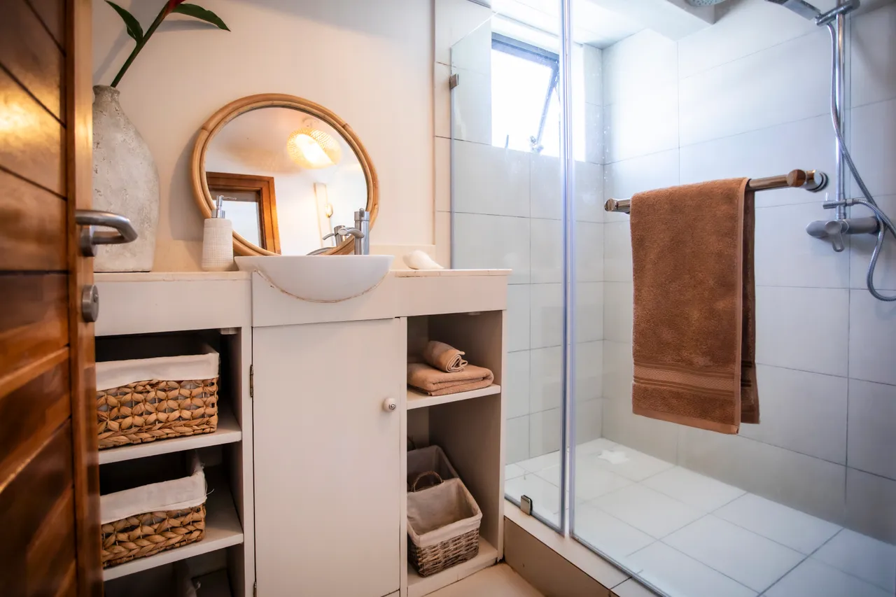 Douche à l'italienne avec parois vitrées, facilitant l'accès et l'entretien.  

Miroir rond encadré de bois pour une touche décorative.  

Rangements ouverts pour serviettes et produits de toilette, favorisant l'organisation.  

Lumière naturelle abondante à travers la fenêtre, créant une ambiance agréable.  

Sol carrelé clair assur