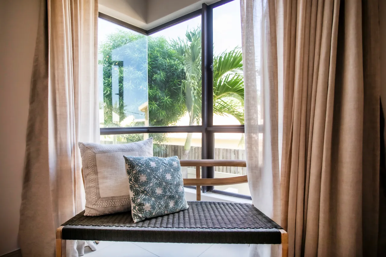 Espace lumineux avec grande fenêtre offrant une vue sur la verdure.  

Banquette confortable agrémentée de coussins pour la détente.  

Rideaux légers permettant à la lumière naturelle d'entrer.  

Matériaux naturels utilisés pour une ambiance apaisante.  

Agencement minimaliste favorisant une atmosphère ouverte et aérée.