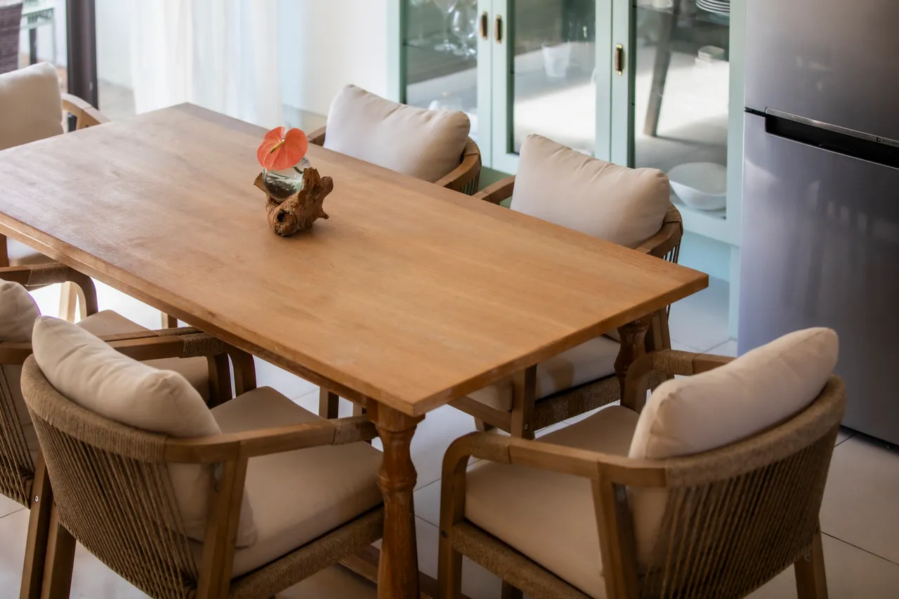 Table en bois entourée de chaises en rotin, offrant un espace convivial.  
Centre de table en bois ajoutant une touche naturelle à l'ensemble.  
Éclairage naturel filtrant par les fenêtres, favorisant une atmosphère sereine.  
Coussins confortables sur les chaises, apportant du confort à l'espace.  
Ambiance calme grâce à un agencement minimaliste