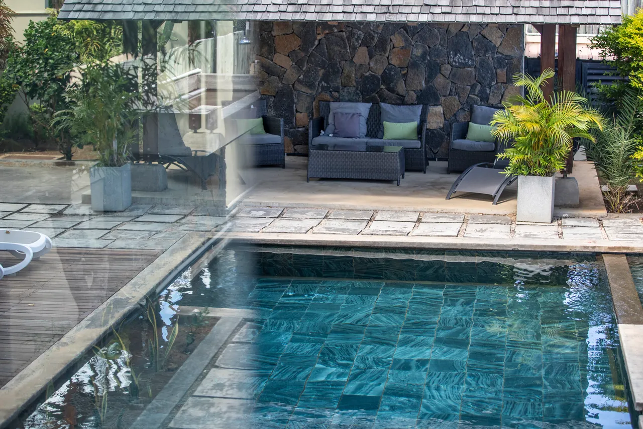 Espace extérieur avec piscine et coin salon ombragé.  
Plantes vertes ajoutant une touche de fraîcheur autour de la piscine.  
Terrasse en pierre bordant l'eau, créant un accès pratique à la piscine.  
Chaises longues disposées pour profiter du soleil à côté de la piscine.  
Mur en pierre apportant un aspect naturel à l'espace extérieur.
