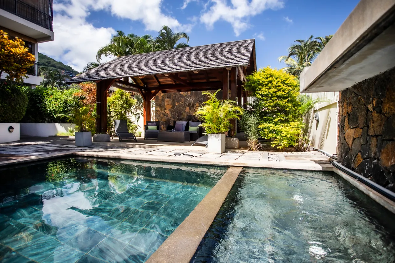 Teintes rafraîchissantes de bleu et vert autour de la piscine.  
Espace ombragé avec mobilier en bois pour se détendre.  
Végétation tropicale offrant un cadre naturel et verdoyant.  
Terrasse en pierre créant un accès direct à la piscine.  
Ambiance calme favorisée par le bruit de l'eau et la verdure environnante.