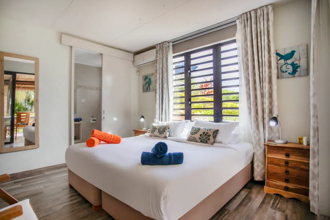 Chambre lumineuse avec lit double et fenêtres à volets. 

Éclairage doux avec lampes de chevet et décor apaisant. 

Rangement pratique avec un bureau en bois intégré. 

Salle de bain attenante équipée d'un miroir et éclairage adapté. 

Couleurs neutres et textile confortable créant une ambiance reposante.