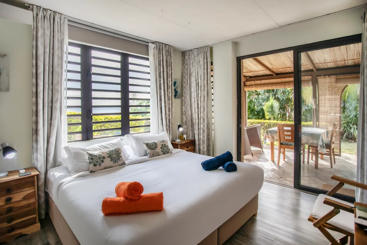 Chambre spacieuse avec lit double et accès direct à l'extérieur. 

Fenêtres à volets offrant une vue sur le jardin. 

Mobilier en bois et décor minimaliste pour une ambiance apaisante. 

Éclairage naturel grâce aux grandes fenêtres et aux rideaux légers. 

Espace de rangement avec des tables de chevet et un bureau intégré.