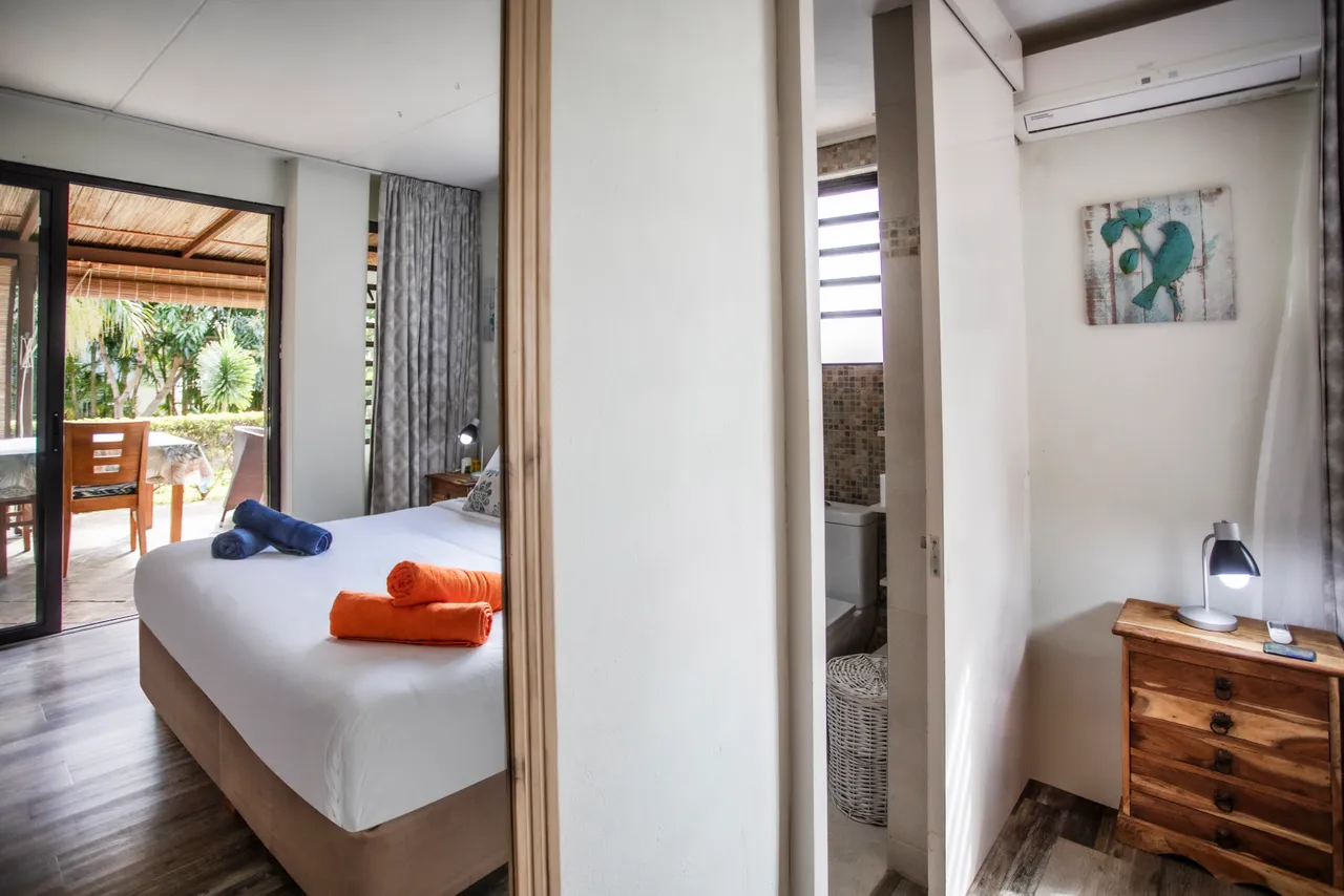 Chambre avec accès à l'extérieur et décor minimaliste. 

Mobilier en bois et rangement intégré pour un espace organisé. 

Éclairage naturel grâce aux grandes fenêtres et rideaux légers. 

Salle de bain attenante avec miroir et coin douche. 

Coussins colorés ajoutant une touche de confort sur le lit.