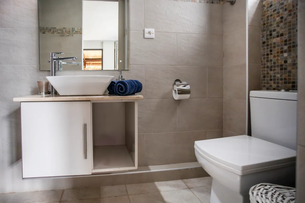 Carrelage clair et murs neutres dans la salle de bain. 

Équipement moderne avec lavabo sur meuble et miroir bien éclairé. 

Serviettes bleues disposées avec soin sur le comptoir. 

Détails de mosaïque ajoutant une touche d'intérêt au mur. 

Espace fonctionnel avec rangement sous le lavabo et toilettes séparées.