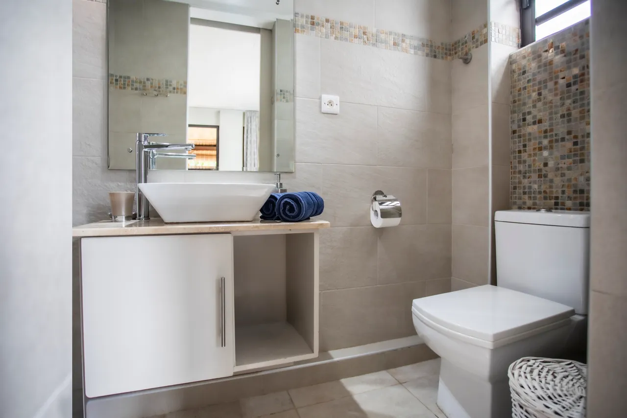 Salle de bain moderne avec carrelage clair et lavabo sur meuble. 

Miroir bien éclairé et rangement sous le comptoir. 

Détails de mosaïque ajoutant une touche colorée au mur. 

Serviettes soigneusement disposées sur le meuble.