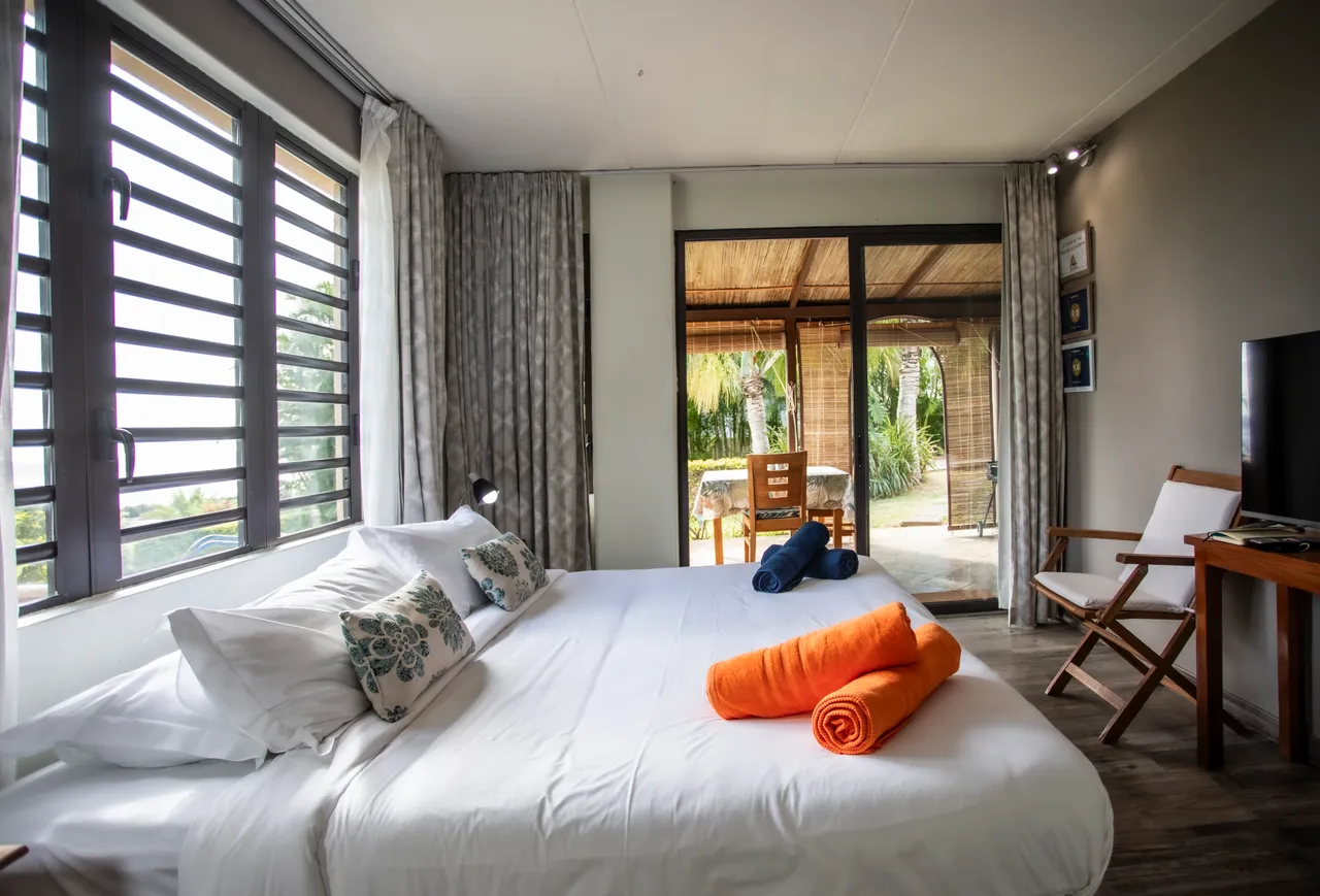 Lit double avec linge de lit blanc et coussins aux motifs floraux.  

Fenêtres à volets avec vue sur l'espace extérieur.  

Mobilier en bois clair et tables de chevet intégrées pour rangement.  

Accès direct à un espace extérieur avec mobilier de jardin.  

Éclairage naturel grâce aux grandes fenêtres et rideaux légers.