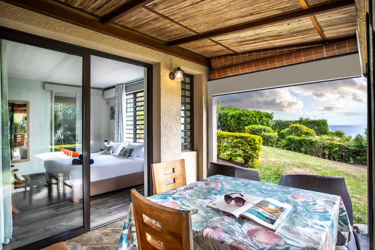 Terrasse couverte avec table et chaises, vue sur le jardin verdoyant.  

Fenêtres coulissantes offrant un accès direct à l'extérieur.  

Mobilier en bois et décor simple pour une atmosphère détendue.  

Éclairage naturel grâce aux grandes ouvertures et aux rideaux légers.