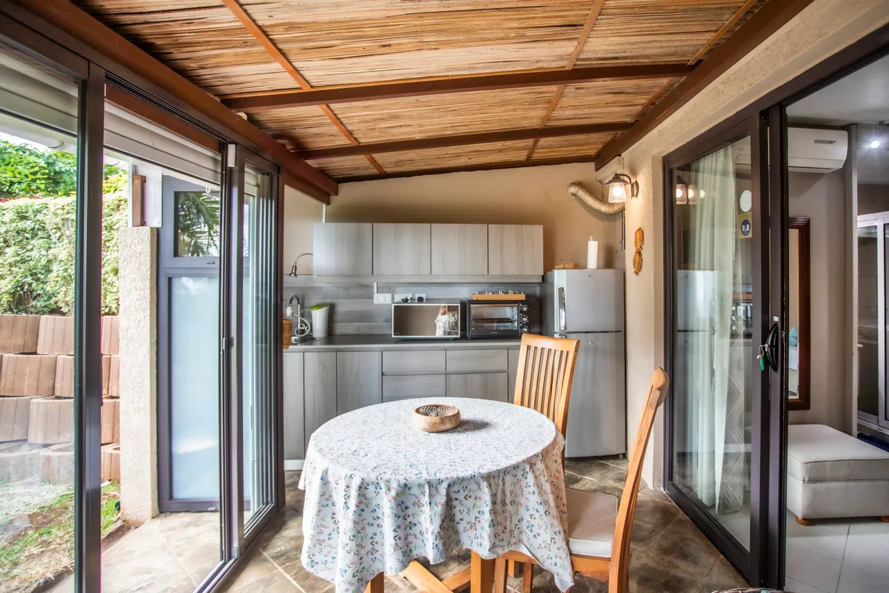 Teintes boisées et claires dans une cuisine ouverte sur la galerie. Coin repas avec table ronde et chaises en bois. Équipement moderne incluant un réfrigérateur et un évier. Lumière naturelle abondante grâce aux grandes fenêtres.