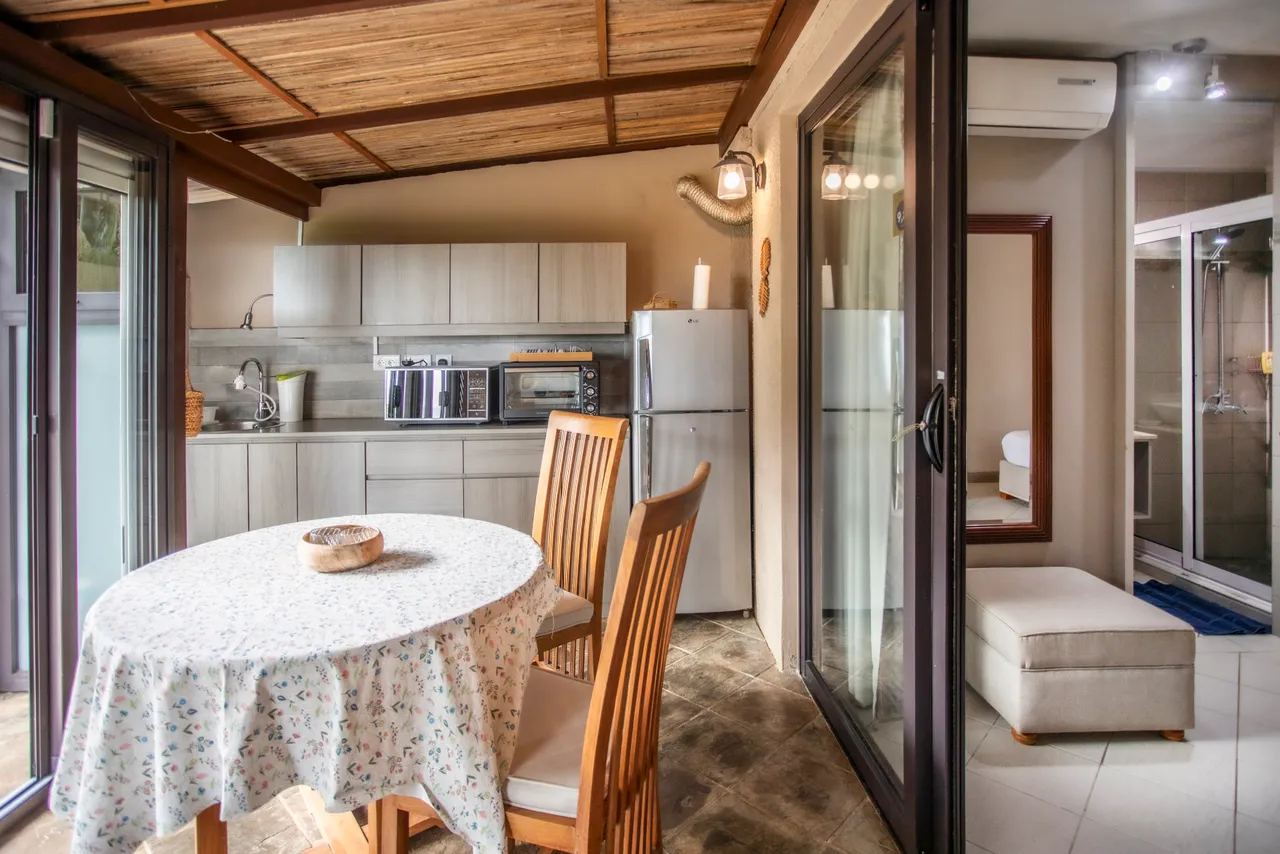 Cuisine avec mobilier clair, équipée d'un réfrigérateur et d'un four à micro-ondes. Coin repas disposant d'une table ronde et de chaises en bois. Lumière naturelle grâce aux grandes fenêtres.