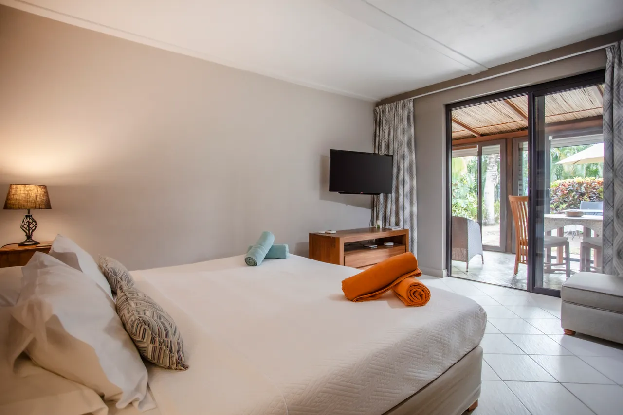 Tons neutres et textures douces dans une chambre avec lit double. Télévision à écran plat et accès à un espace extérieur. Draps et serviettes disposés sur le lit. Espace de rangement intégré et éclairage doux.