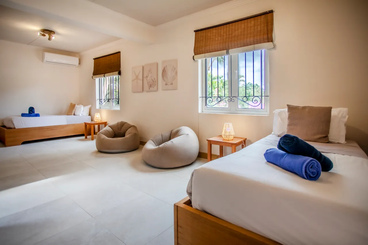 Lumière naturelle à travers les fenêtres, créant une ambiance apaisante. Chambre équipée de deux lits et de poufs confortables. Décor minimaliste avec des touches de couleur sur le linge de lit. Accès direct à la vue sur l'extérieur depuis les fenêtres.