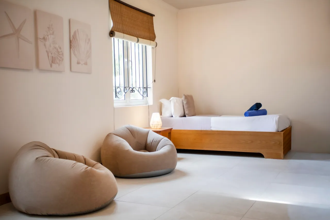 Chambre avec un lit simple et deux poufs en tissu. Décor sobre avec des œuvres murales.