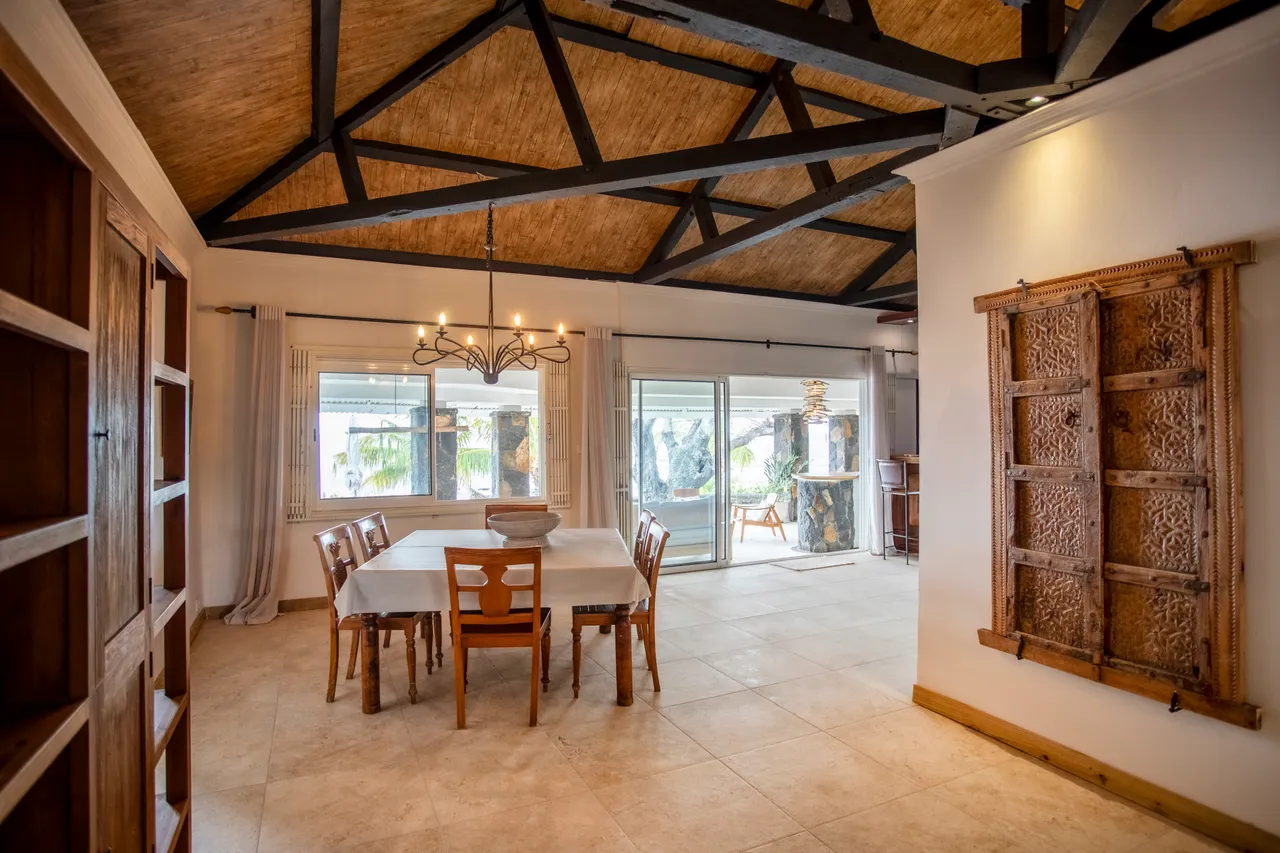 Espace repas avec une grande table en bois et chaises en bois clair. Plafond à poutres apparentes et éclairage suspendu. Grandes baies vitrées offrant une vue sur l'extérieur.