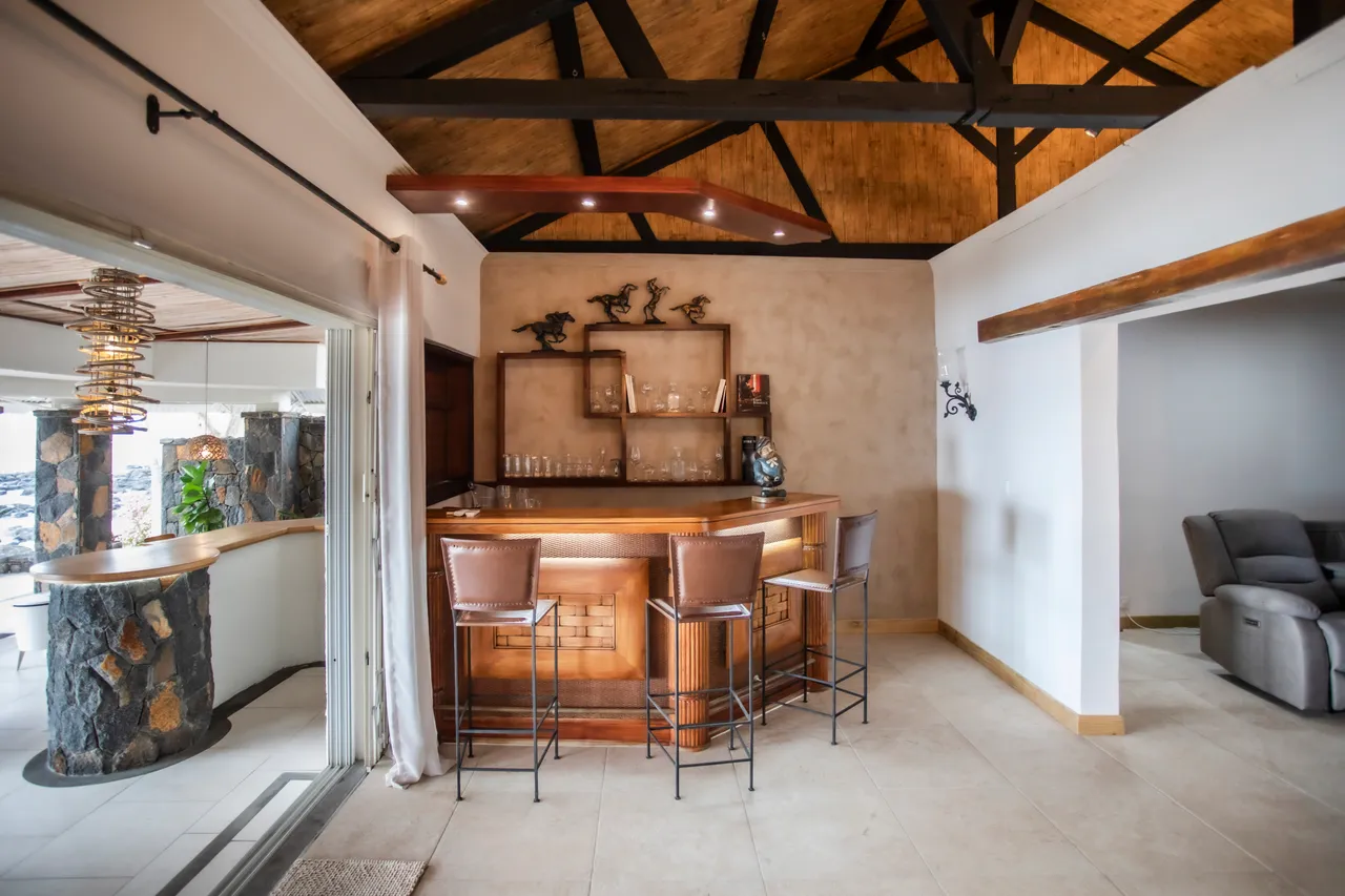 Espace repas avec un bar en bois et des tabourets assortis. Plafond à poutres noires et éclairage intégré. Accès direct à la terrasse extérieure.