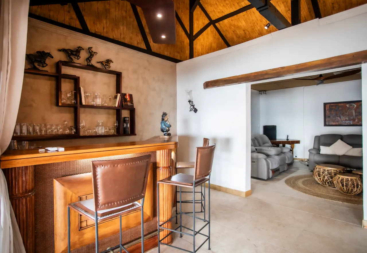 Teintes chaudes du bois enrichissent l'espace bar. Tabourets en cuir face à un comptoir en bois. Éclairage discret accentuant le plafond haut. Décoration épurée avec œuvres murales en fond.