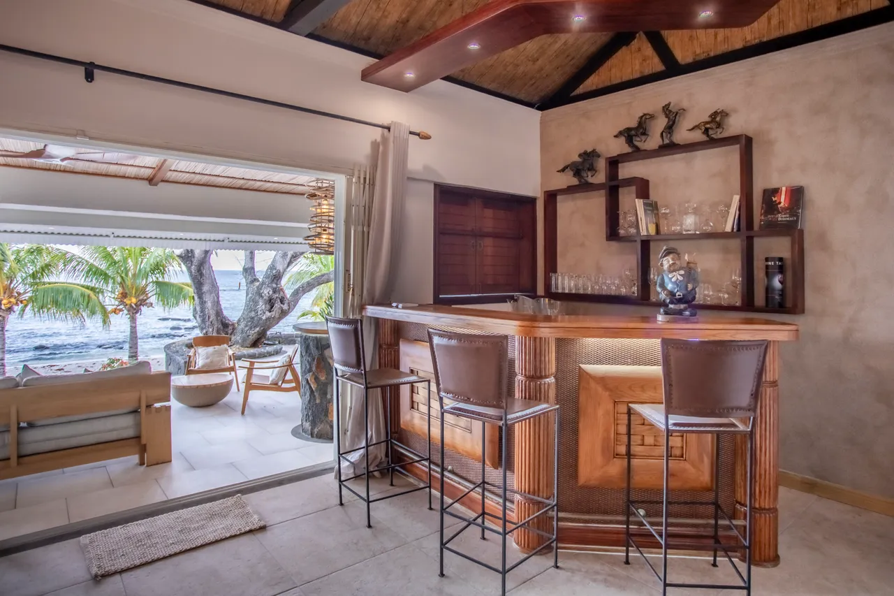 Espace repas avec un bar en bois et quatre tabourets en cuir. Ouverture sur la terrasse et vue sur la mer. Éclairage intégré au plafond en bois. Décor sobre et moderne avec éléments en verre.