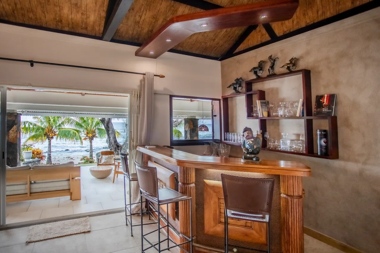 Bois sombre du bar en harmonie avec le plafond à poutres. Éclairage discret mettant en valeur l'espace et la vue. Terrasse accessible depuis l'espace, avec vue sur la mer.