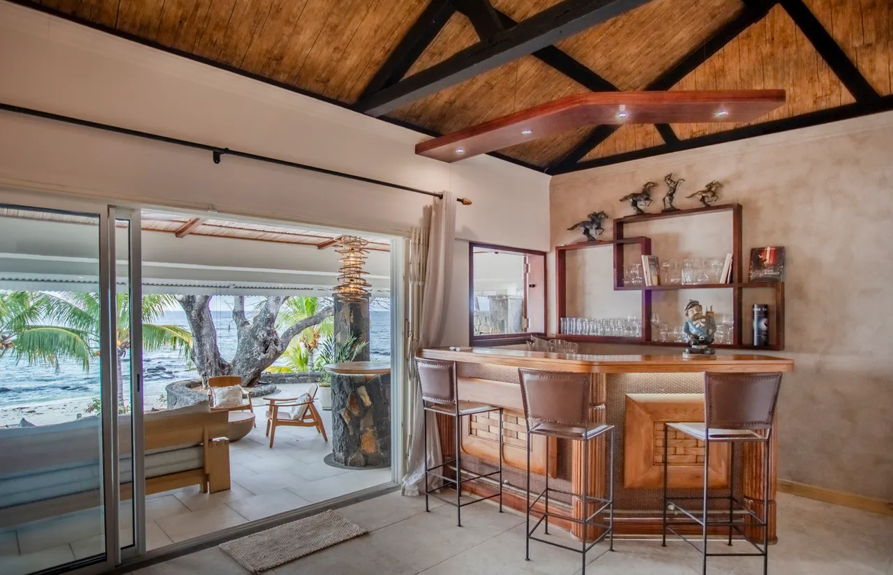 Bar en bois avec tabourets, accès direct à la terrasse. Vue sur la mer et les palmiers.