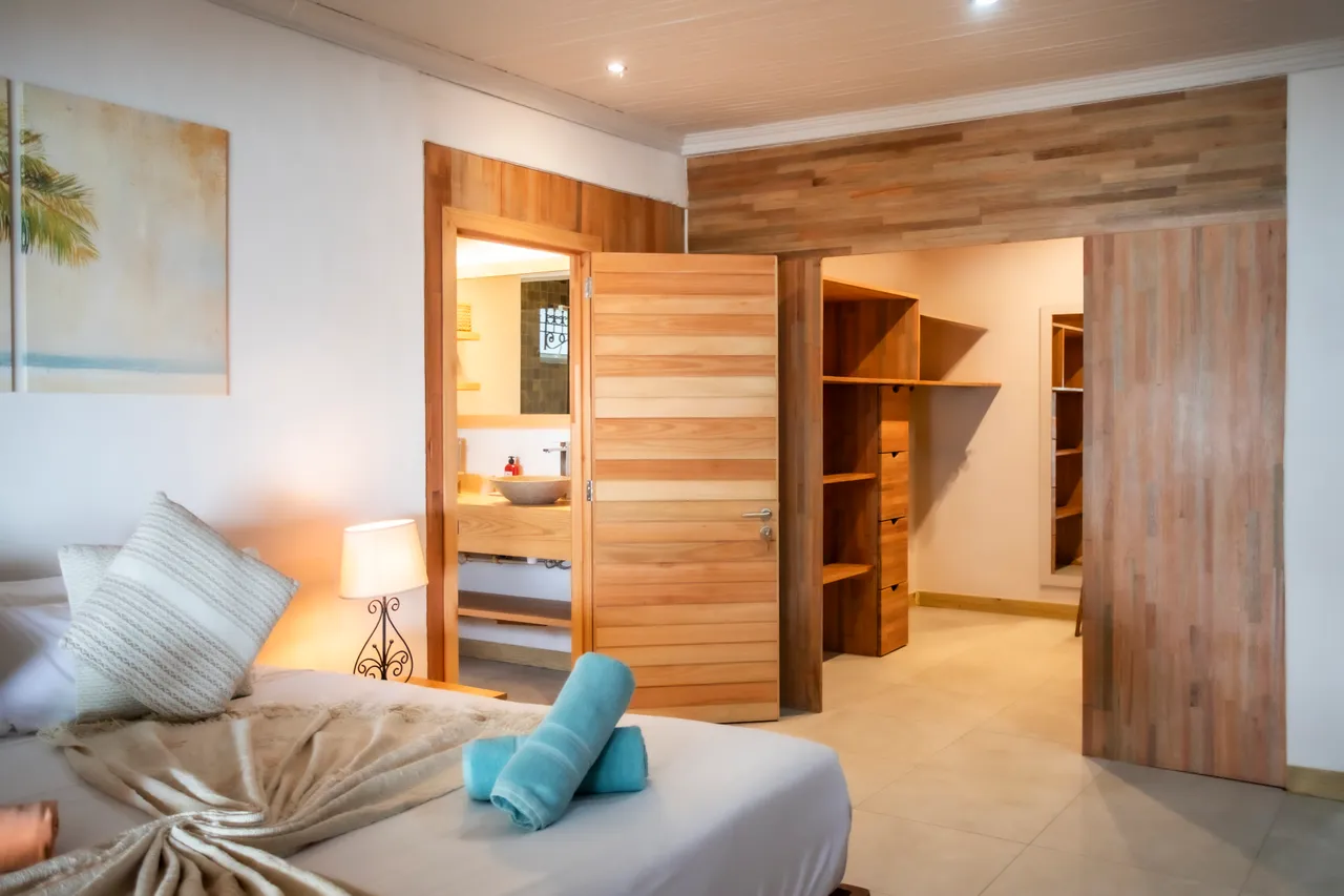 Chambre avec un lit double, tables de chevet et accès à un dressing. Éclairage doux et rangements intégrés. Salle de bain attenante visible à l'arrière.