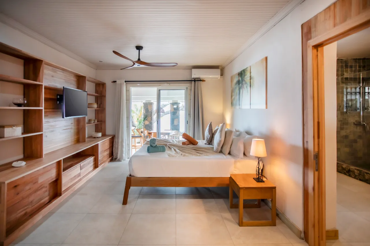 Chambre avec un lit double et un meuble télé en bois. Fenêtres coulissantes donnant sur la terrasse. Éclairage subtil grâce aux lampes de chevet et au plafond. Salle de bain attenante avec douche moderne en mosaïque.