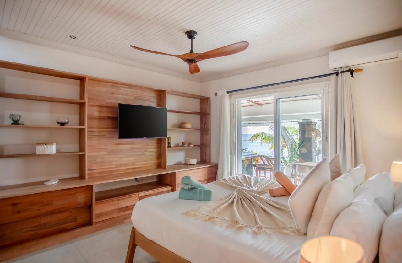Chambre avec lit double et mobilier en bois. Accès à une terrasse vue mer. Éclairage naturel et télévision murale.