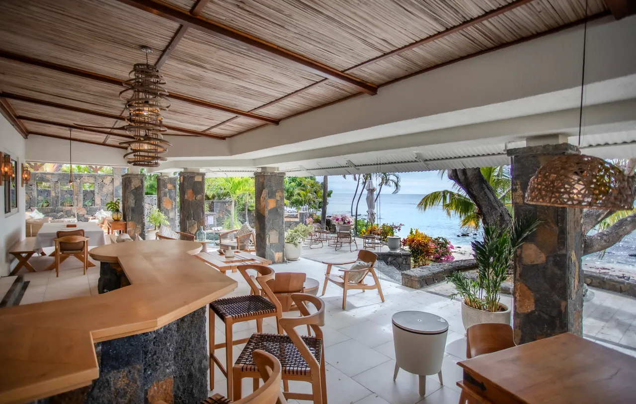 Le bois naturel du bar s'intègre harmonieusement au décor. Espace ouvert avec vue sur la mer et accès à la terrasse. Mobilier en bois et éclairage tamisé créent une ambiance accueillante.