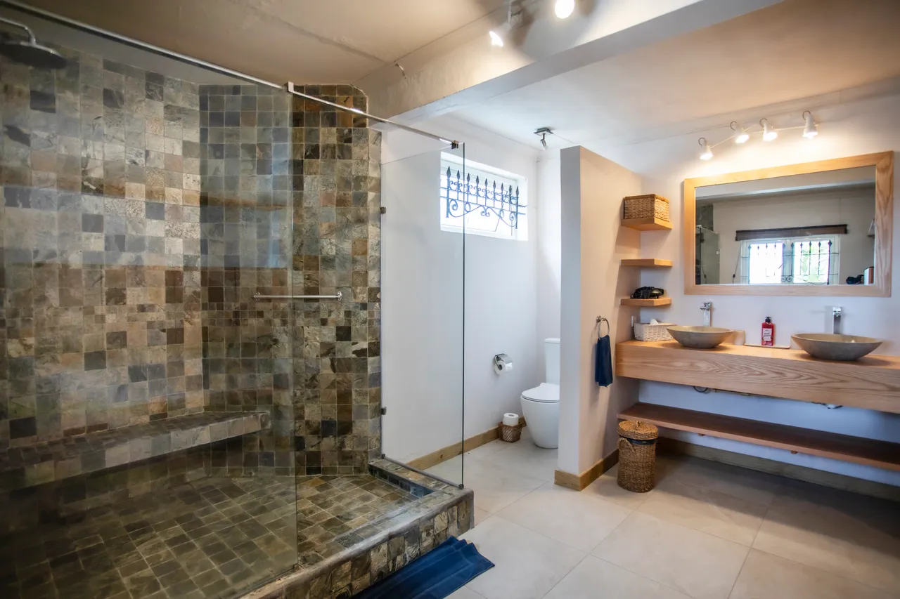 Texture pierreuse avec carrelage en mosaïque dans la douche. Lavabo en bois avec vasques modernes. Miroir large apportant de la luminosité à l'espace. Étagères en bois pour un rangement pratique.