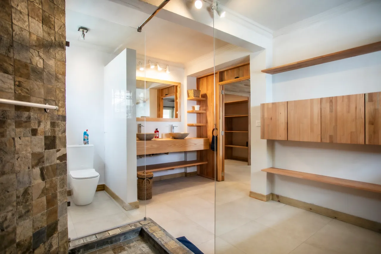 Salle de bain avec double vasque et miroir éclairant. Douche carrelée avec séparation en verre. Étagères en bois pour un rangement efficace.