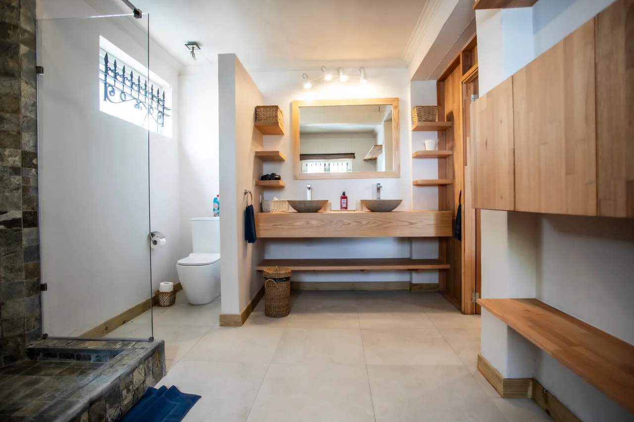 Salle de bain avec carrelage clair et large miroir lumineux. Étagères en bois pour un rangement fonctionnel. Douche avec paroi en verre et éclairage intégré.