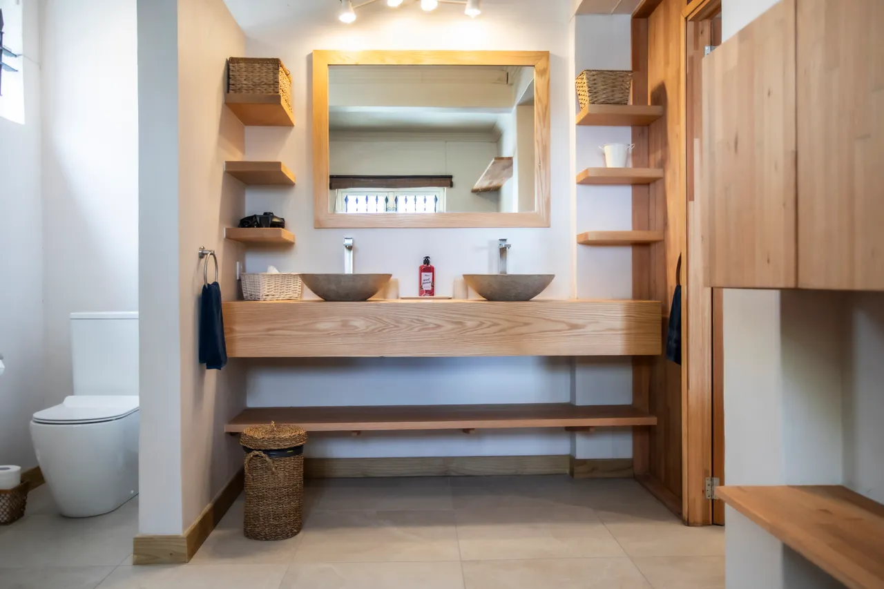 Salle de bain avec double vasque en pierre et étagères en bois. Éclairage naturel et miroir amplifiant l’espace.