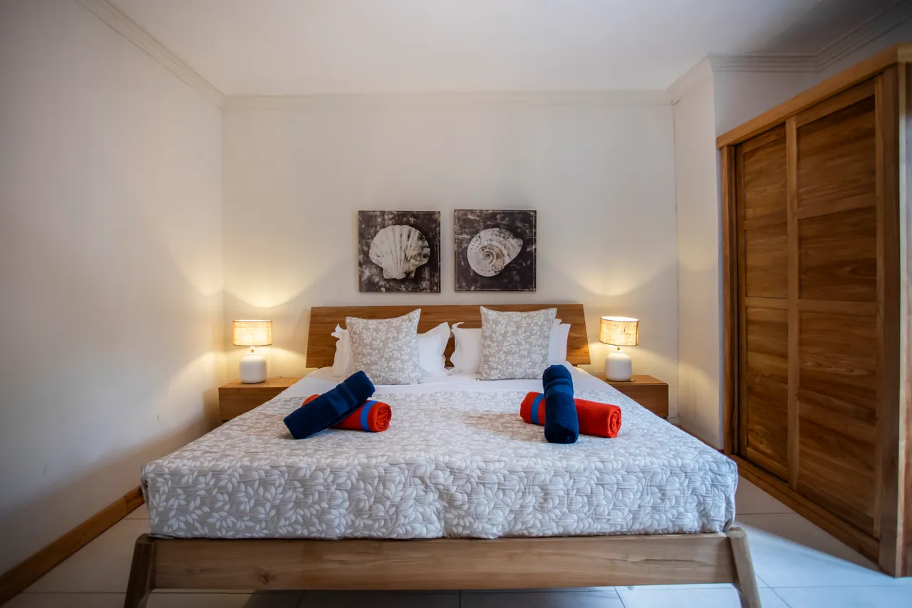 Chambre lumineuse avec lit en bois et linge de lit clair. Deux lampes de chevet apportent une ambiance douce. Rangement pratique avec un placard intégré en bois.