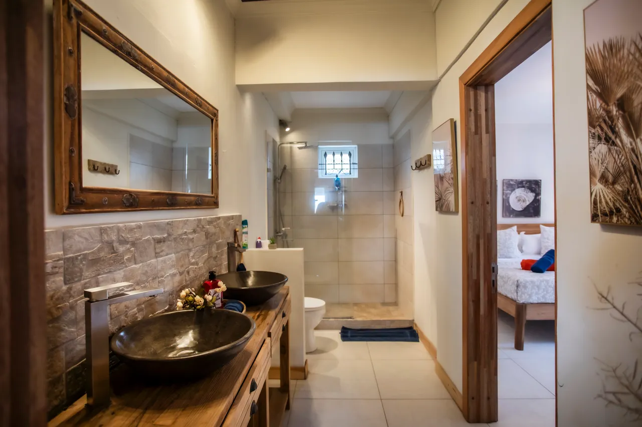 Couleurs neutres avec carrelage clair et grandes douches. Deux vasques en pierre et miroir encadré. Étagères en bois pour un rangement optimisé. Douche avec paroi en verre et éclairage intégré.