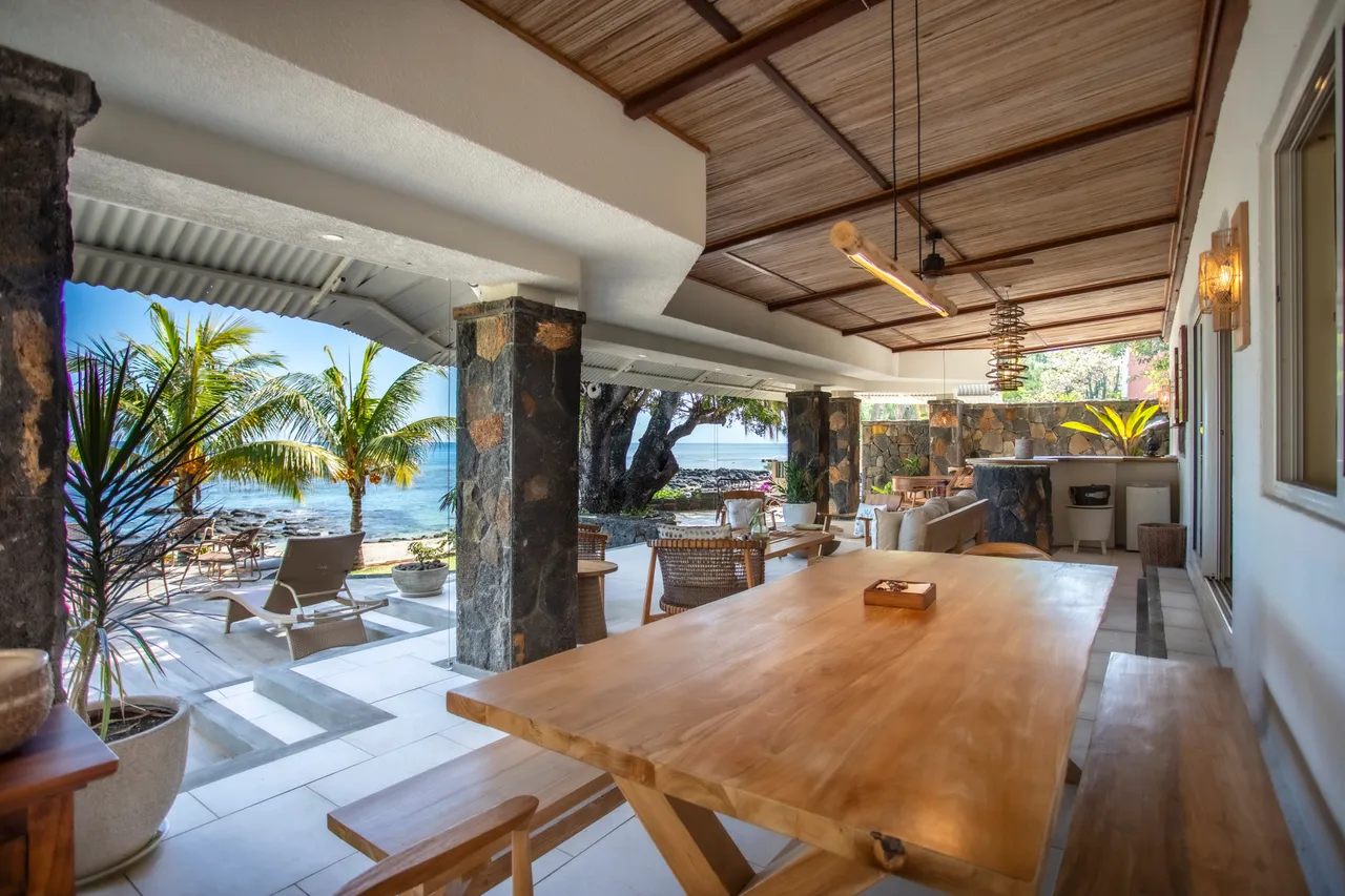 Espace repas extérieur avec grande table en bois et vue sur la mer. Palmiers ajoutent une touche naturelle.