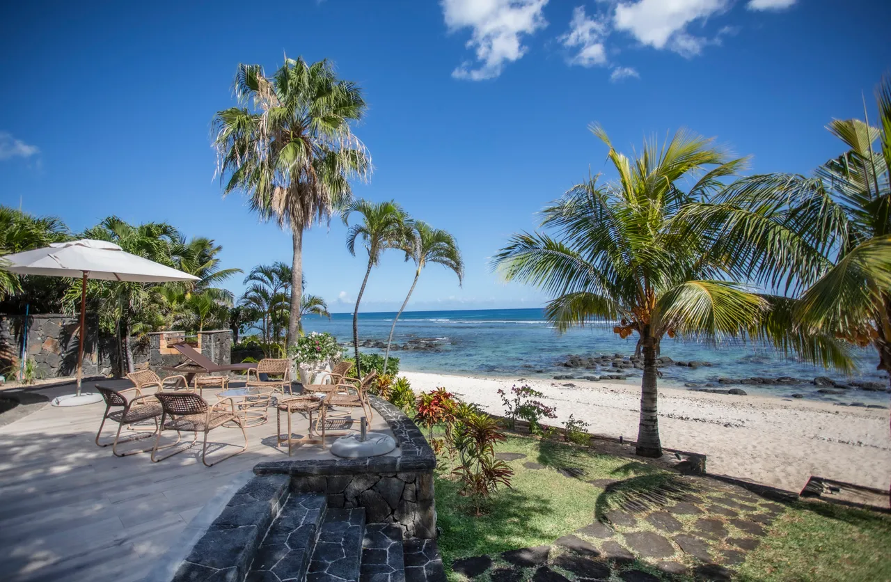 Terrasse extérieure avec mobilier en rotin, vue sur la mer et la plage. Palmiers ajoutent une touche tropicale.